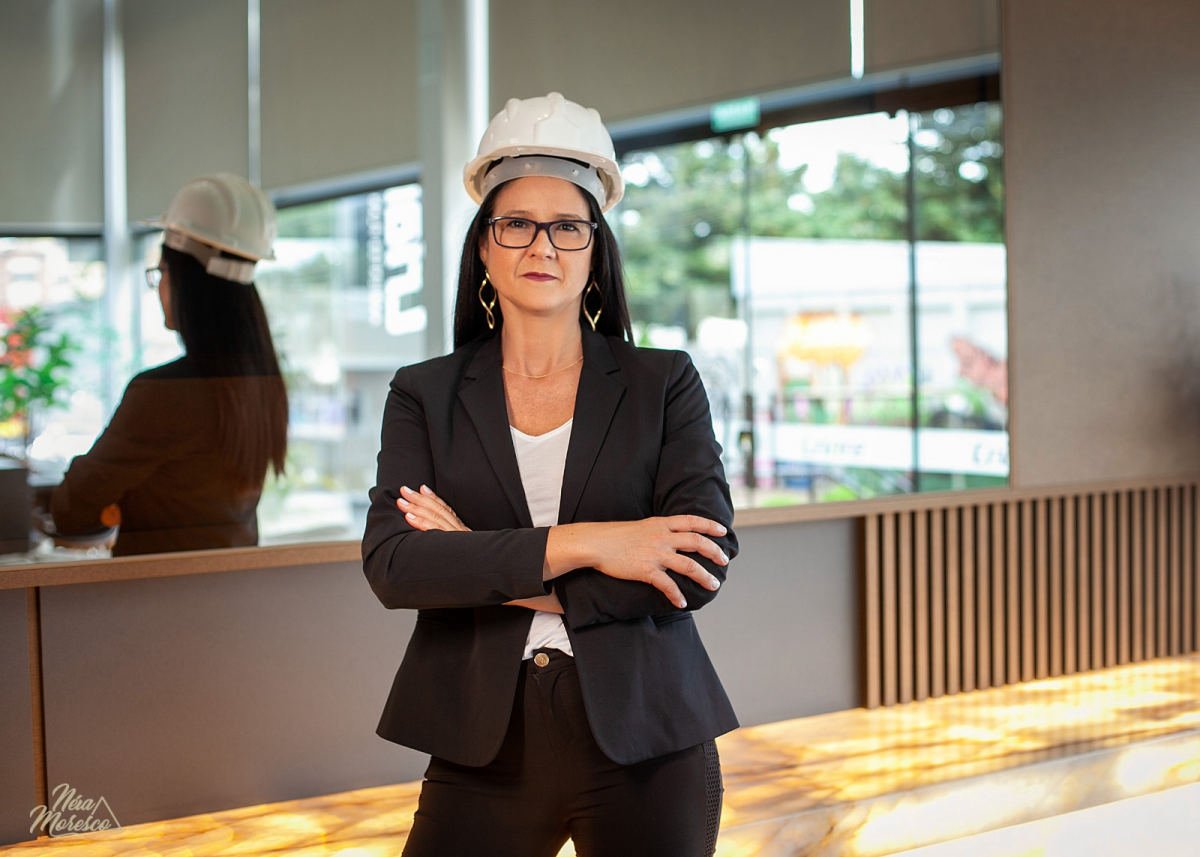 Marta Rejane Tessaro de braços cruzados, com capacete branco e blazer, no hall de um empreendimento